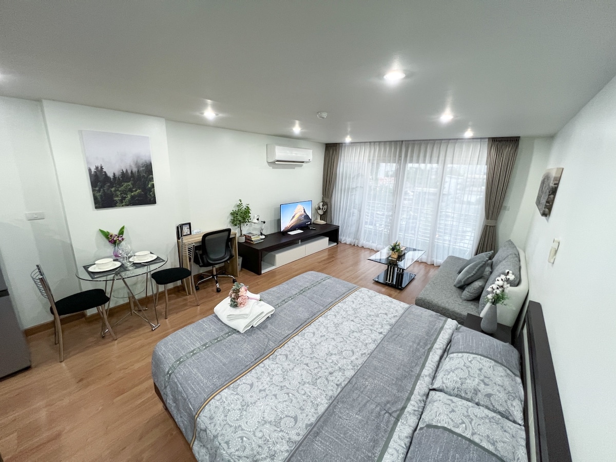 The spacious room features a large bed with decorative pillows, complemented by a stylish area with a couch and a coffee table. A dining table with black chairs is positioned nearby. Natural light enters through expansive windows covered with sheer curtains, illuminating the contemporary decor.