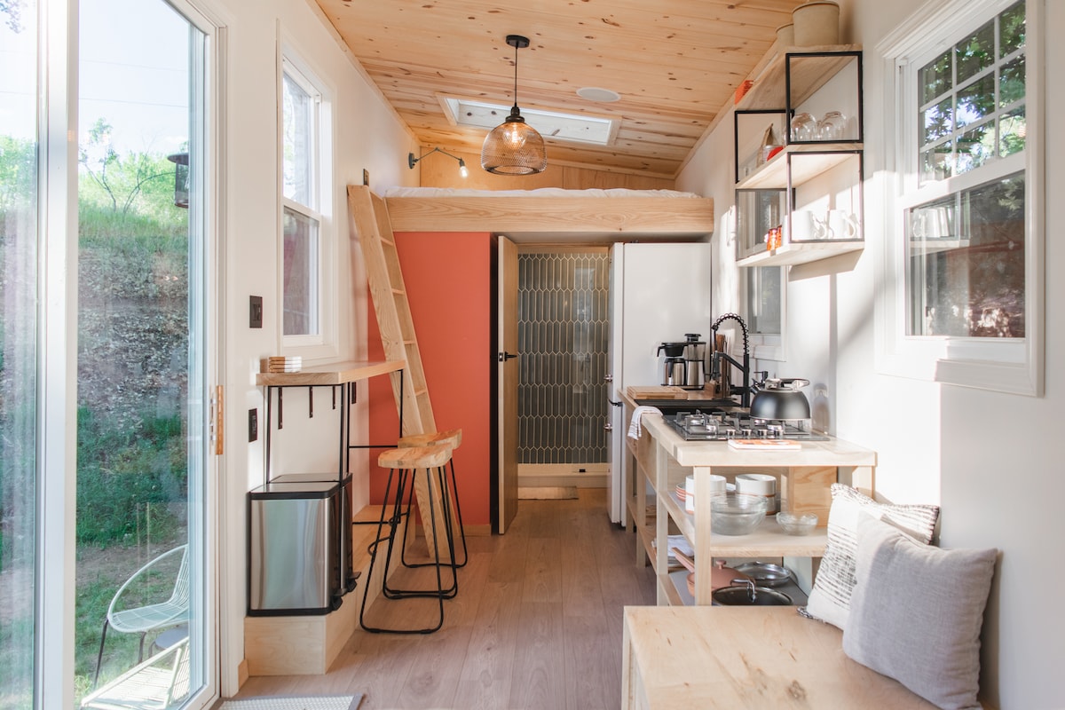The open kitchen area features light wooden cabinetry and modern appliances, including a refrigerator and coffeemaker. A breakfast bar with two stools provides a casual dining space. Natural light streams in through large windows, illuminating the minimalist decor.