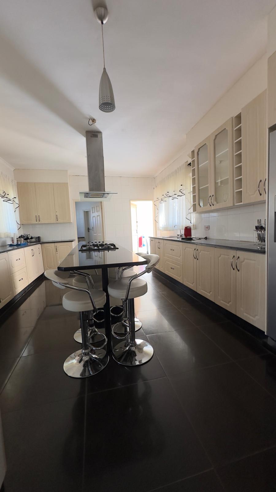 A spacious kitchen is presented with sleek black countertops and light-colored cabinetry. Modern bar stools surround a central island, with ample storage evident in cabinets and shelves. Natural light filters in through multiple windows, creating a bright and functional cooking space.