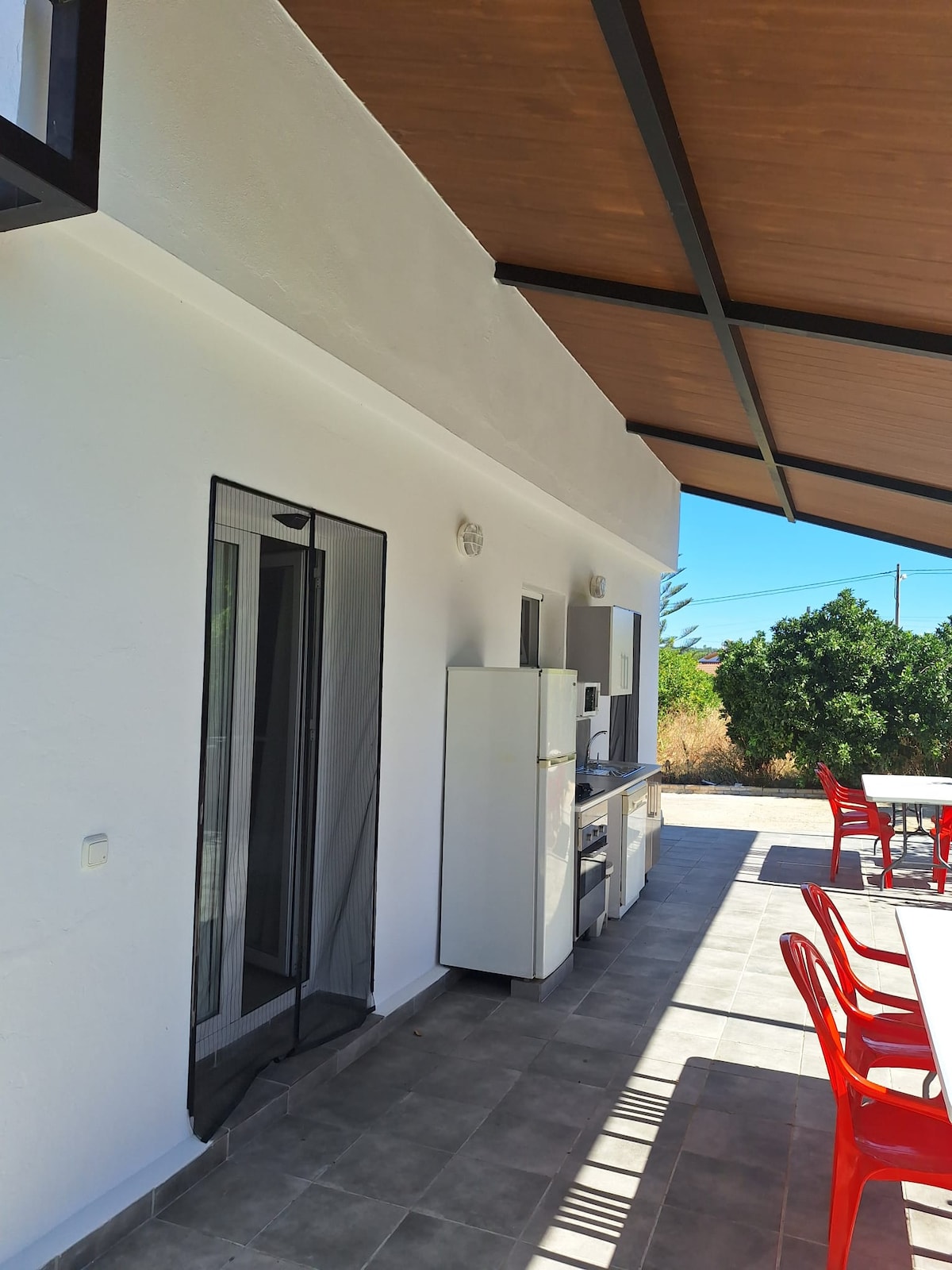 An outdoor area is shown, featuring a spacious porch with a tiled floor underneath a sloping roof. A shared kitchen setup can be seen along one wall, including a refrigerator and cooking appliances. Several red chairs surround a table, providing an inviting space for guests.
