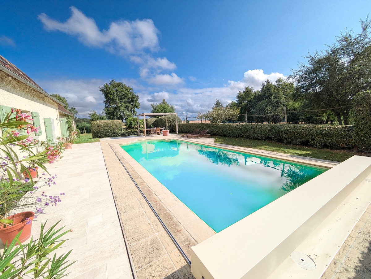An inviting outdoor space features a large swimming pool surrounded by sunbathing areas. Lush greenery and hedges create a private ambiance, while the clear blue sky adds brightness. Comfortable seating under a shaded pergola is present, inviting relaxation and enjoyment in the tranquil surroundings.