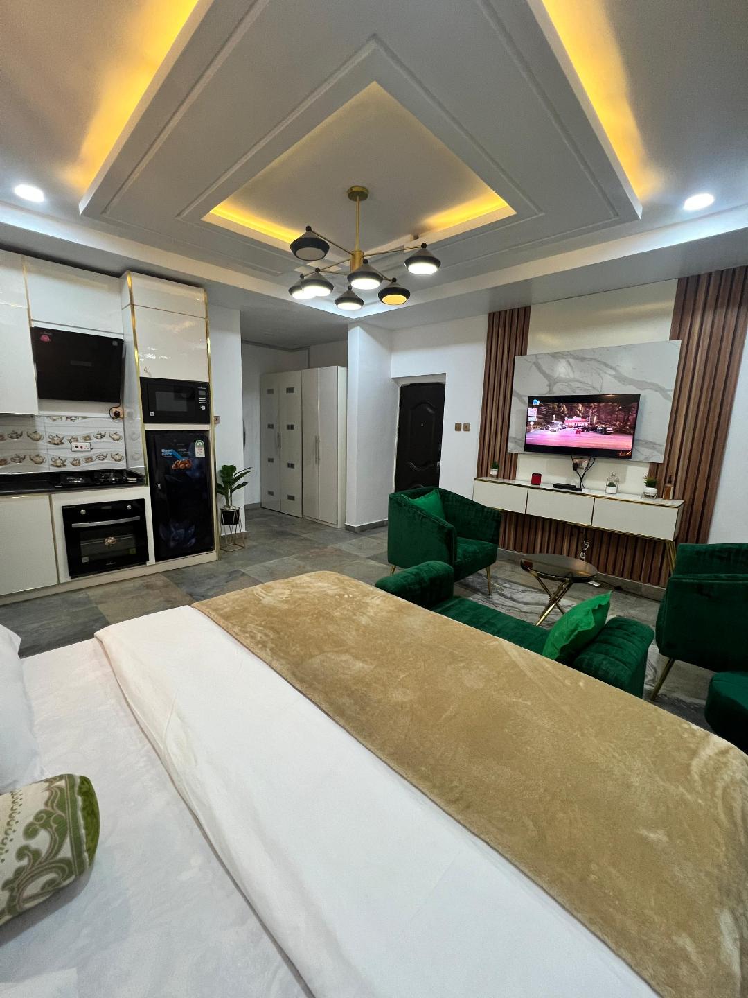 The studio apartment features a modern kitchenette equipped with essential appliances. A cozy sleeping area is situated next to a stylish entertainment setup, which includes a flat-screen SMART TV. Green accent chairs provide additional seating against the tastefully designed backdrop.