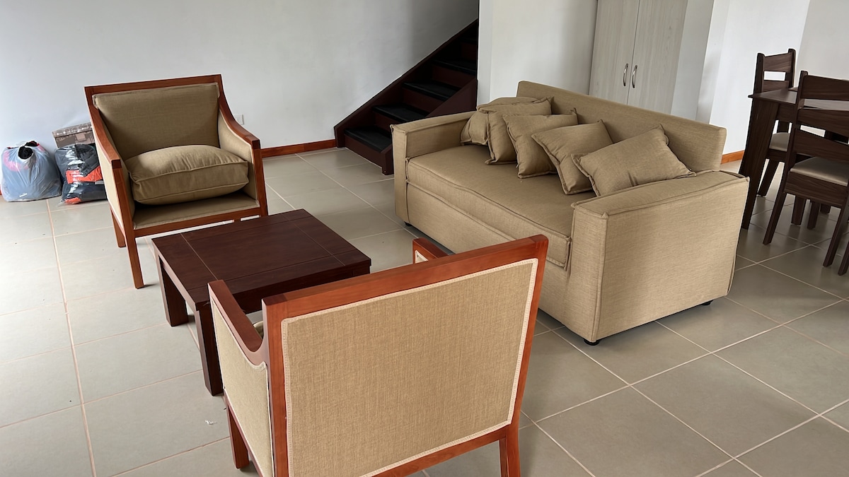 A living area is presented with a comfortable sofa adorned with several cushions, accompanied by two armchairs and a wooden coffee table. A dining table can be seen in the background, and a staircase is visible to one side.