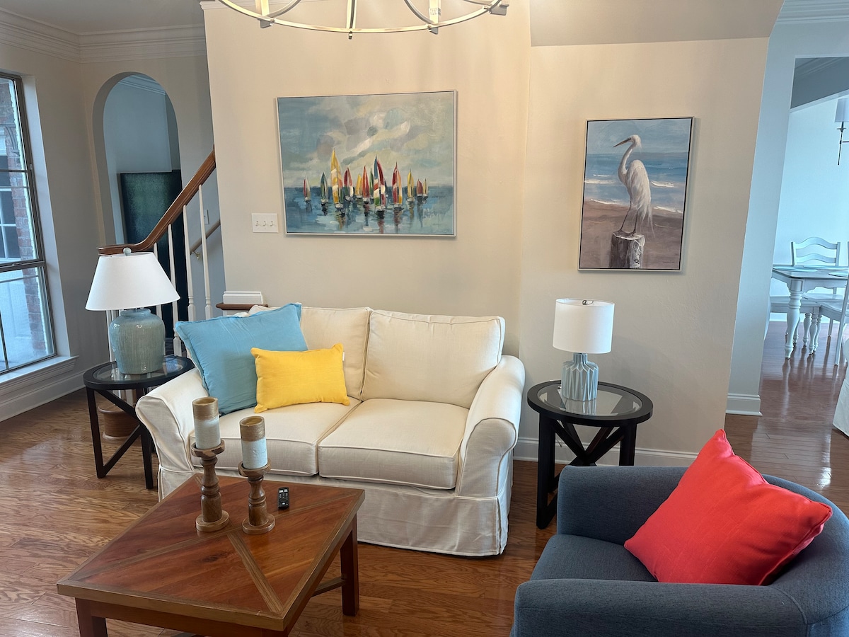 A living area features a light-colored sofa adorned with colorful throw pillows, accompanied by a blue armchair. A wooden coffee table is centered beneath a chandelier. Walls are embellished with framed artwork depicting boats and wildlife. Natural light enters through a large window.