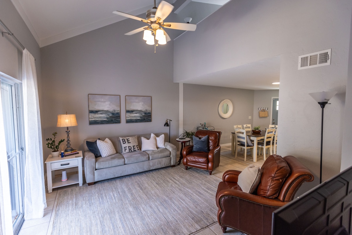 A bright living area is depicted with a light grey color scheme. A cozy sofa and a leather chair are invitingly arranged around a textured rug. Two framed artworks adorn the wall, and a dining area is visible in the background, illuminated by natural light.