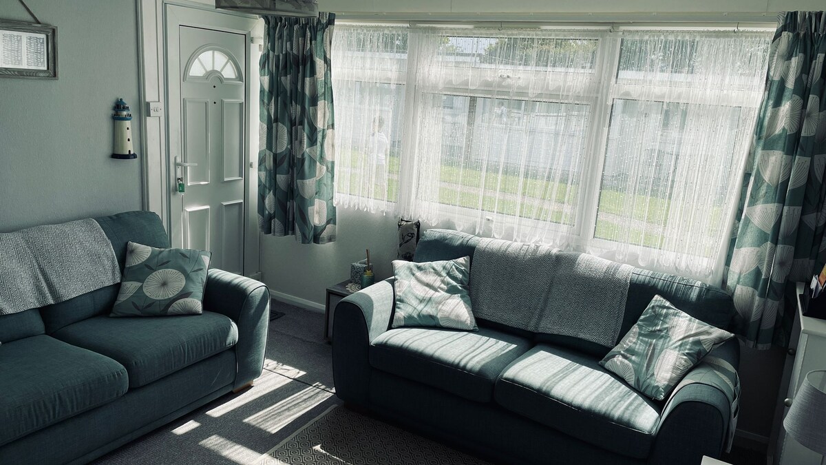 A bright living area is characterized by large windows dressed with sheer curtains, allowing natural light to fill the space. Two comfortable sofas in muted teal tones are positioned invitingly, accompanied by decorative cushions. The entry door features a windowpane design, enhancing light and openness.