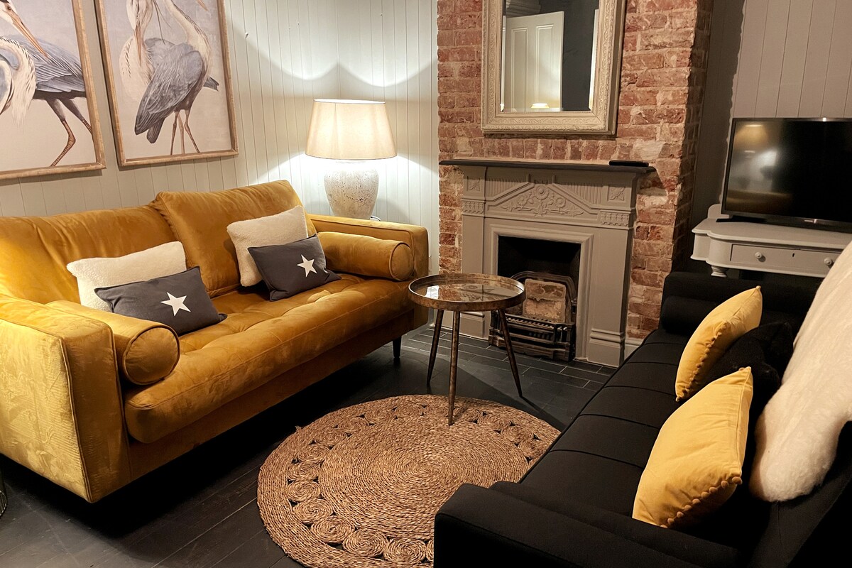 The sitting room features a vibrant yellow sofa and a dark, stylish couch, arranged around a circular glass-topped table. A soft area rug lies beneath, while artwork adorns the walls. A traditional fireplace and a flat-screen television complete the cozy ambiance.