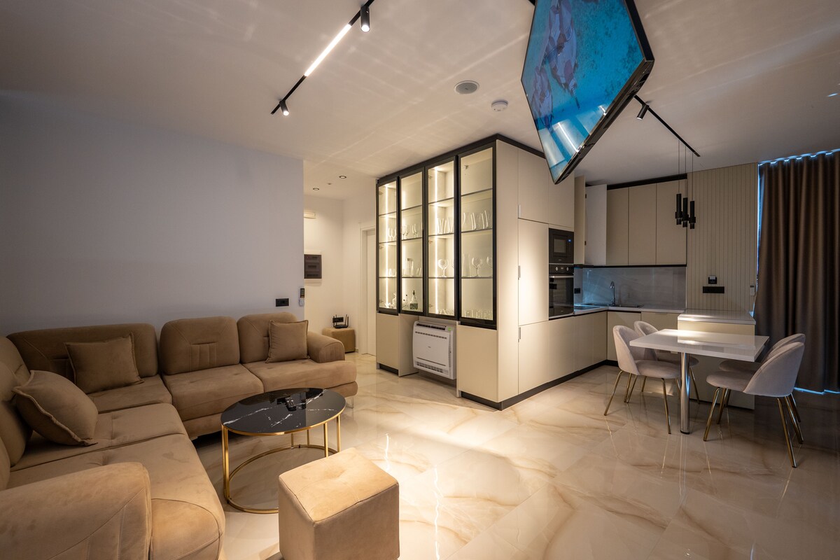 A spacious living area is presented with a sectional sofa in neutral tones, a circular glass coffee table, and an elegant dining table with upholstered chairs. A large flat-screen TV is mounted above a sleek kitchen, which features modern appliances and cabinetry. Natural light is enhanced by large windows.