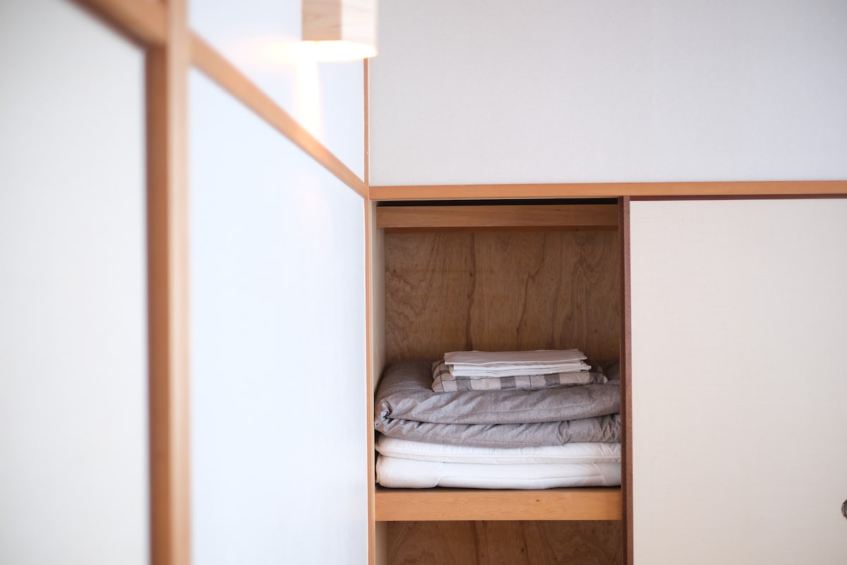 A neatly organized shelf displays stacks of bedding in neutral tones. Soft linens and blankets are arranged methodically, showcasing a tidy and inviting space. The warm wooden frame of the shelf adds a touch of natural elegance.