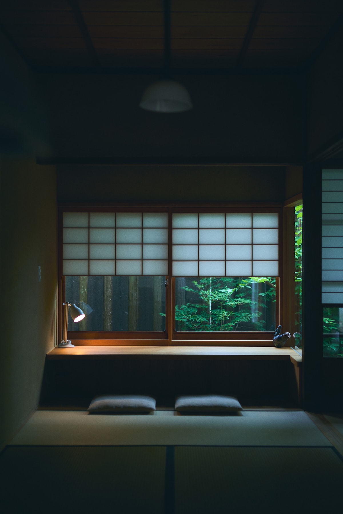 A serene corner of the room features a wooden window with shoji screens, allowing soft light to filter through. A small desk with a lamp and two cushions are arranged below, providing a peaceful spot to enjoy views of the lush greenery outside.