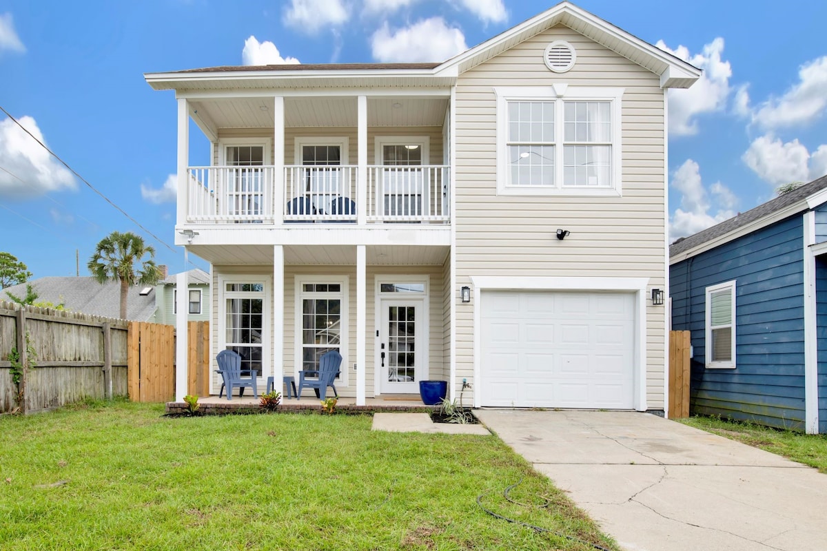 The exterior of a newly remodeled home is showcased, featuring a light-colored siding, a private garage, and a spacious yard. Two blue chairs are located on the porch, complemented by potted plants. Bright white windows reflect natural light, enhancing the welcoming appearance of the property.