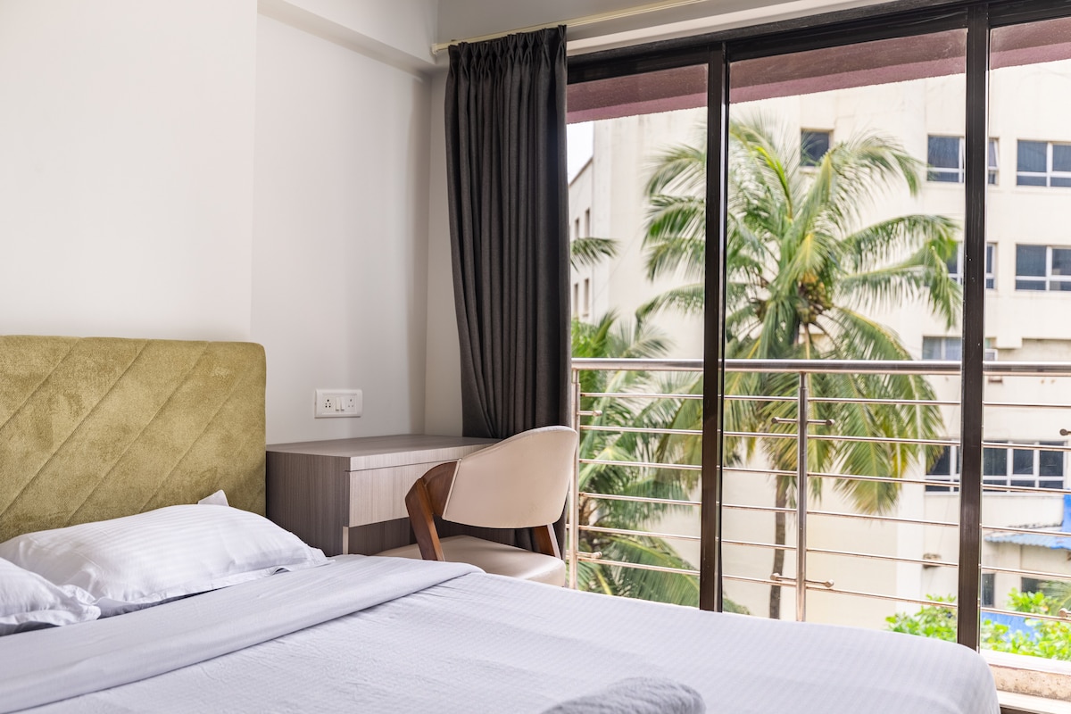 A bright bedroom features a queen-size bed with crisp linens, accompanied by a modern chair and desk. Large sliding glass doors provide natural light and views of palm trees outside, while dark curtains create a contrast with the soft, neutral tones of the room.