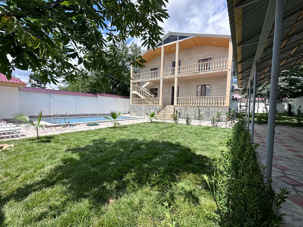 A well-maintained outdoor space features a large grassy area bordered by low shrubs. A private swimming pool is visible, accompanied by a spacious two-story building with balconies. The scene is shaded by trees, offering a serene environment for relaxation.