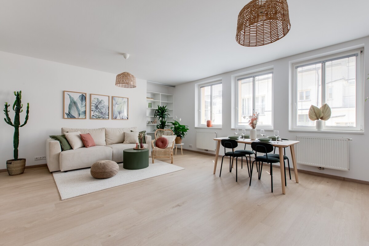 A light and airy living space features a spacious sofa adorned with decorative pillows. A natural fiber rug anchors the seating area. A dining table is set for four, surrounded by sleek chairs. Large windows allow ample natural light, highlighting the greenery of potted plants.