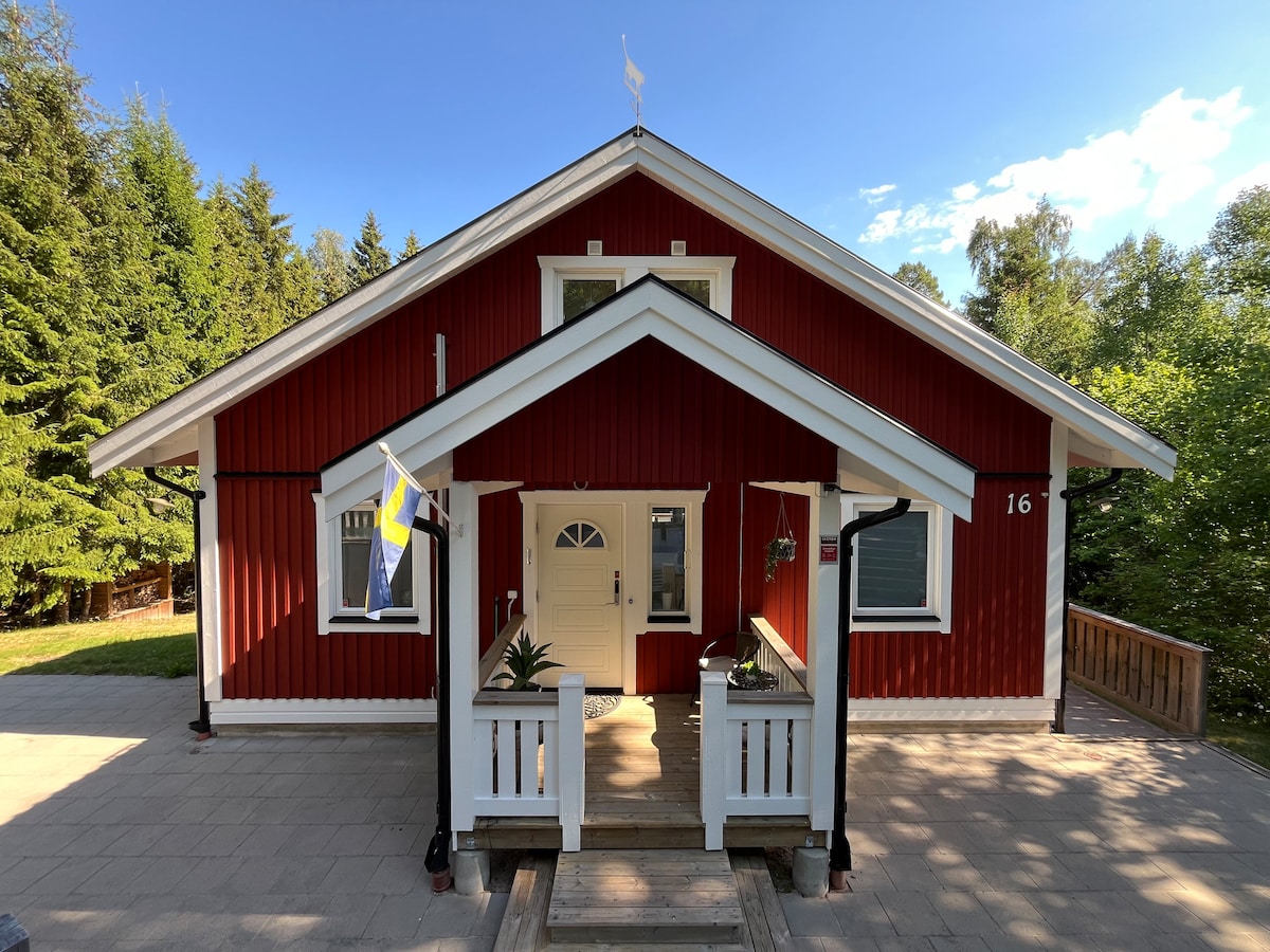 A charming red cottage is framed by lush green trees. The entrance features a welcoming porch with white railings and steps leading up to the door. A small flag can be seen, adding a touch of character to the inviting facade.