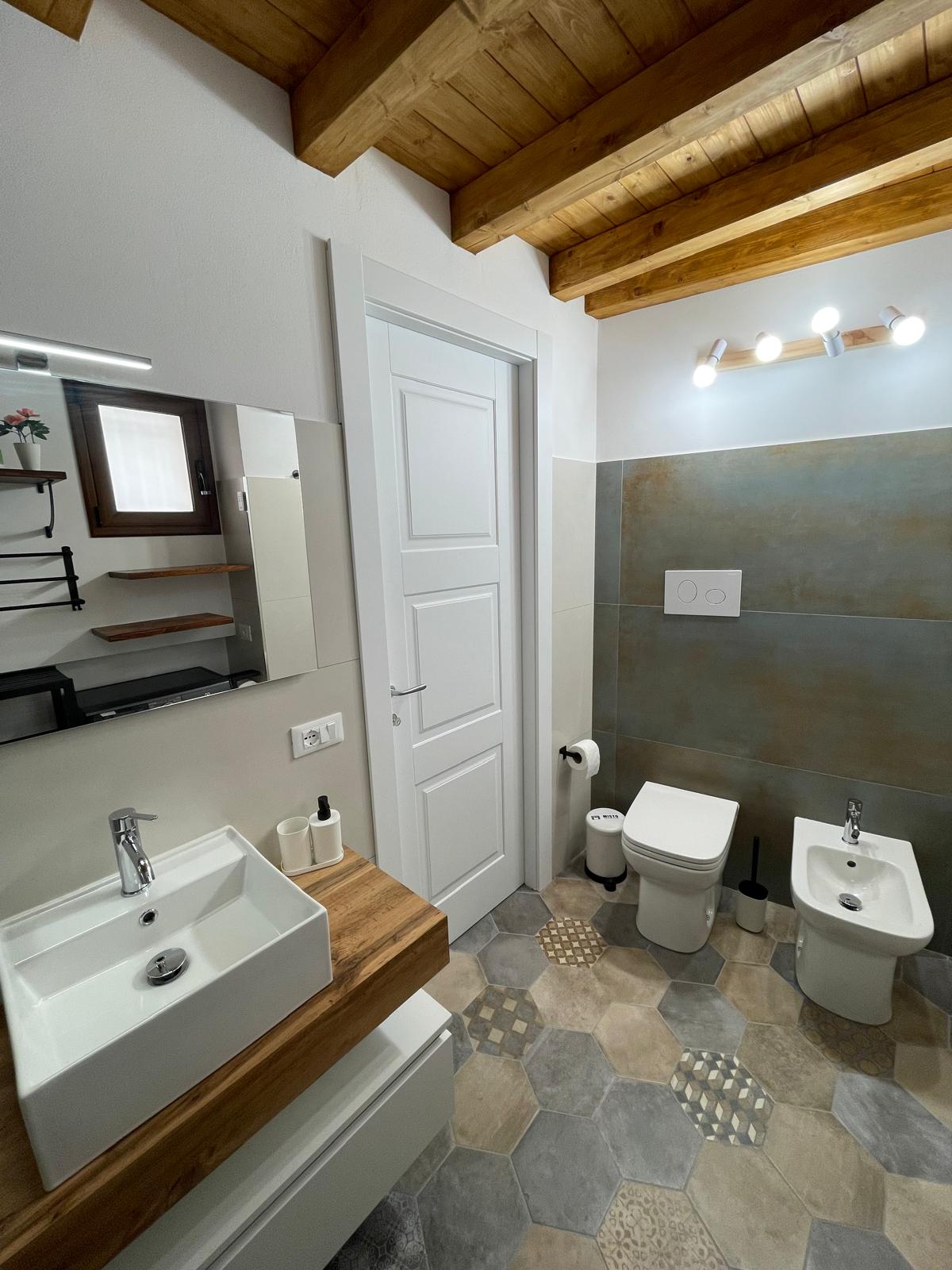 The bathroom features a modern design with a wooden ceiling and neutral tile flooring. A sleek vanity with a square sink is complemented by a large mirror. A toilet and bidet are situated beside each other, with wall-mounted shelves providing additional storage.