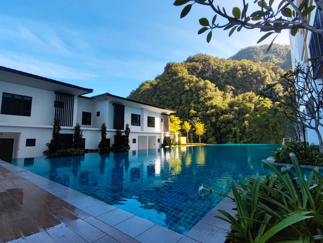 An expansive infinity pool reflects the clear blue sky, surrounded by lush greenery and gentle slopes of the hillside. Modern buildings with large windows are positioned nearby, enhancing the tranquil environment. The pool area is complemented by neatly landscaped plants at the forefront.