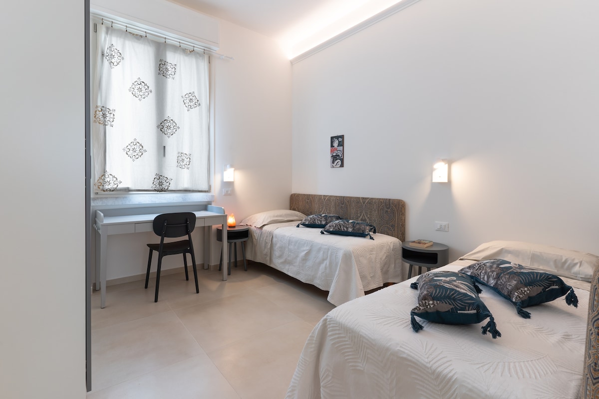 A well-lit room features two single beds, each adorned with soft linens. A small desk with a chair is positioned near a window dressed with a patterned curtain. Wall sconces provide gentle illumination, enhancing the room's functional layout.