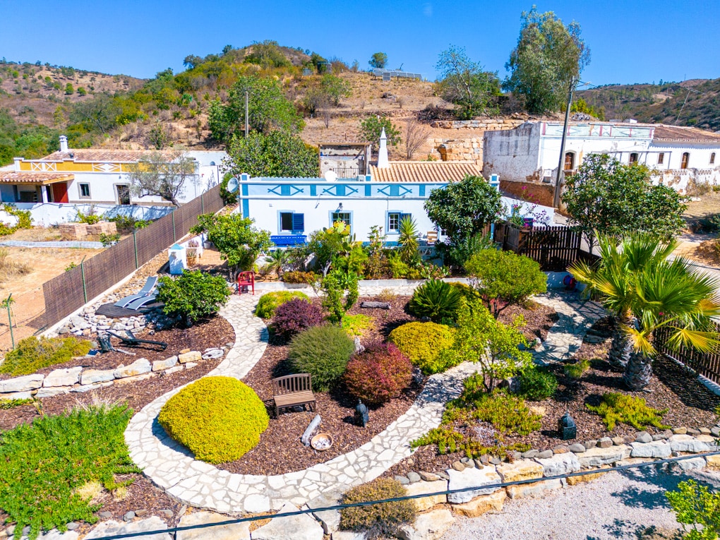 The outdoor space features a landscaped garden with a winding stone pathway, vibrant greenery, and assorted plants. A bench is placed amidst the foliage, providing a serene spot for relaxation. The surrounding hills add a natural backdrop to this peaceful countryside setting.