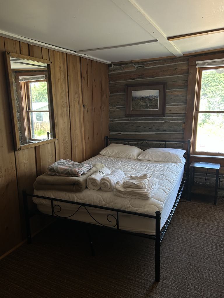A private room features rustic log walls and large windows allowing natural light. A full-size bed is neatly made with linens, flanked by two windows. A mirror and a framed artwork add to the simple decor. Towels are neatly stacked on the bed.