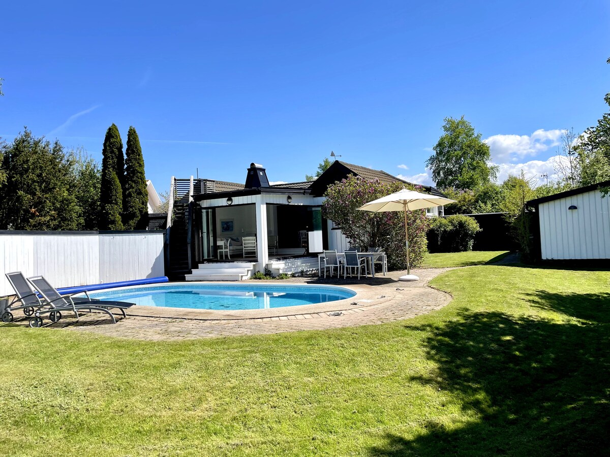 A well-maintained garden showcases a swimming pool surrounded by a spacious patio. Lawn chairs are positioned near the pool, and a large umbrella provides shade. In the background, a house with large windows offers a bright, inviting ambiance.