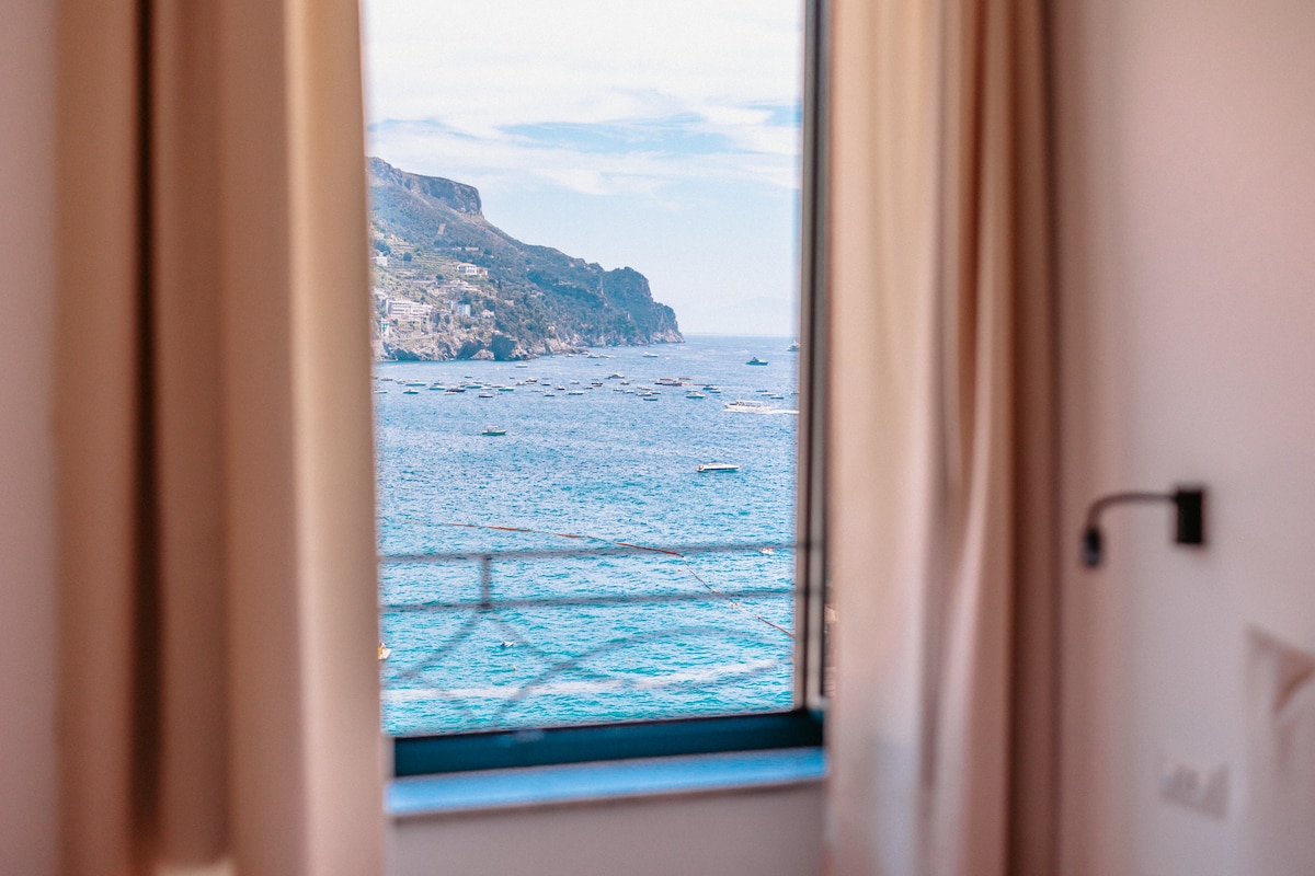 A scenic view of the ocean is framed by light, flowing curtains. The horizon features gentle waves, with distant boats dotting the water. Soft natural light filters through the window, enhancing the serene atmosphere of the space.