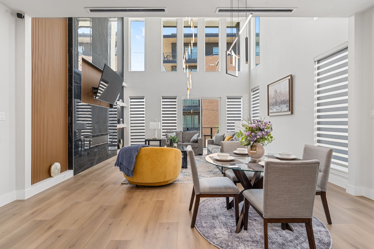The open living area features a large dining table surrounded by upholstered chairs. A modern sofa sits alongside a wall-mounted TV. Natural light floods through multiple windows, highlighting the stylish decor and use of clean lines throughout the space.