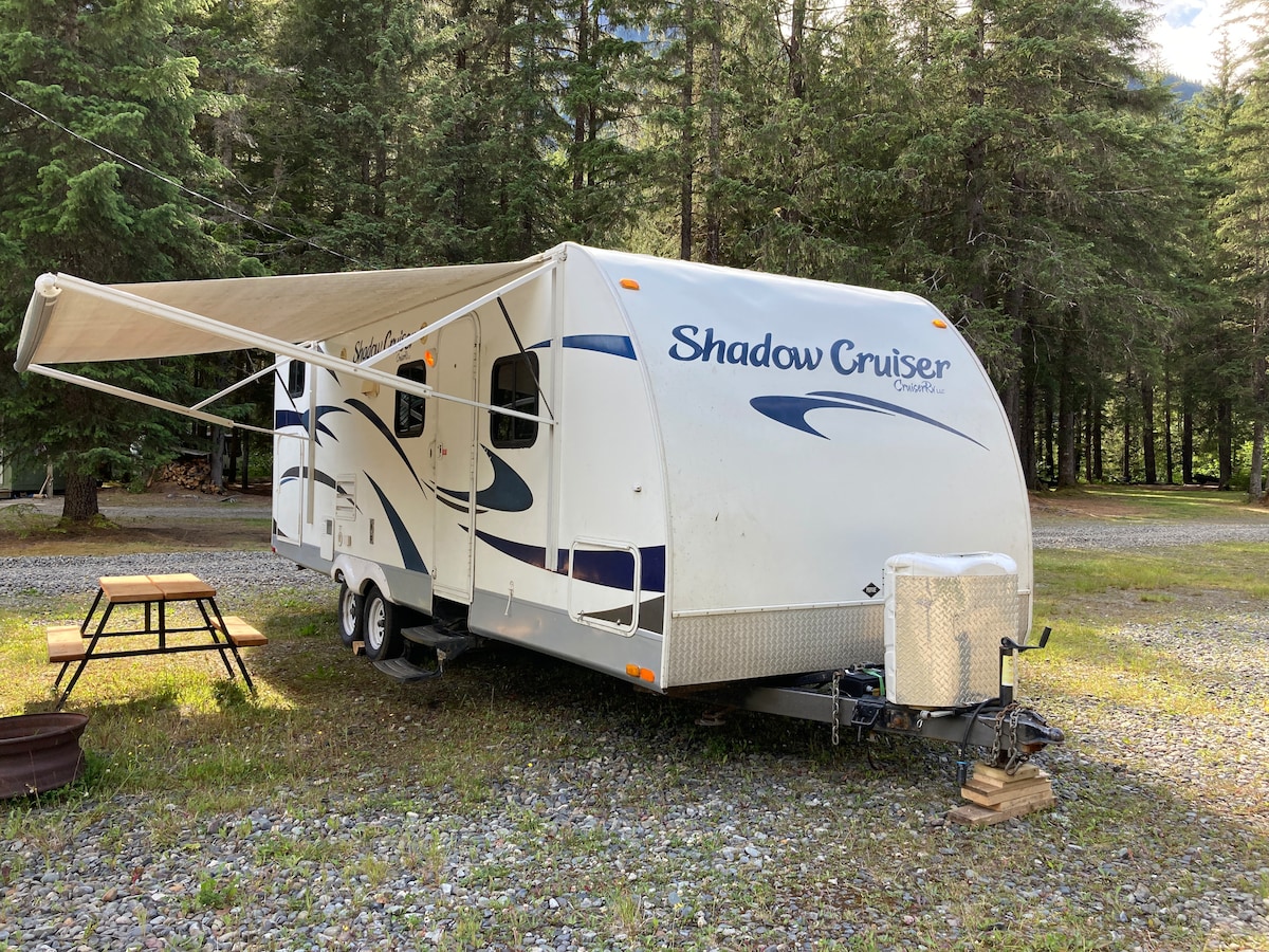 The stationary RV camper, featuring a streamlined design, is parked on a gravel site surrounded by tall, lush trees. An extended awning provides shade over a picnic table nearby, while a fire pit is situated on the ground, enhancing the outdoor experience.