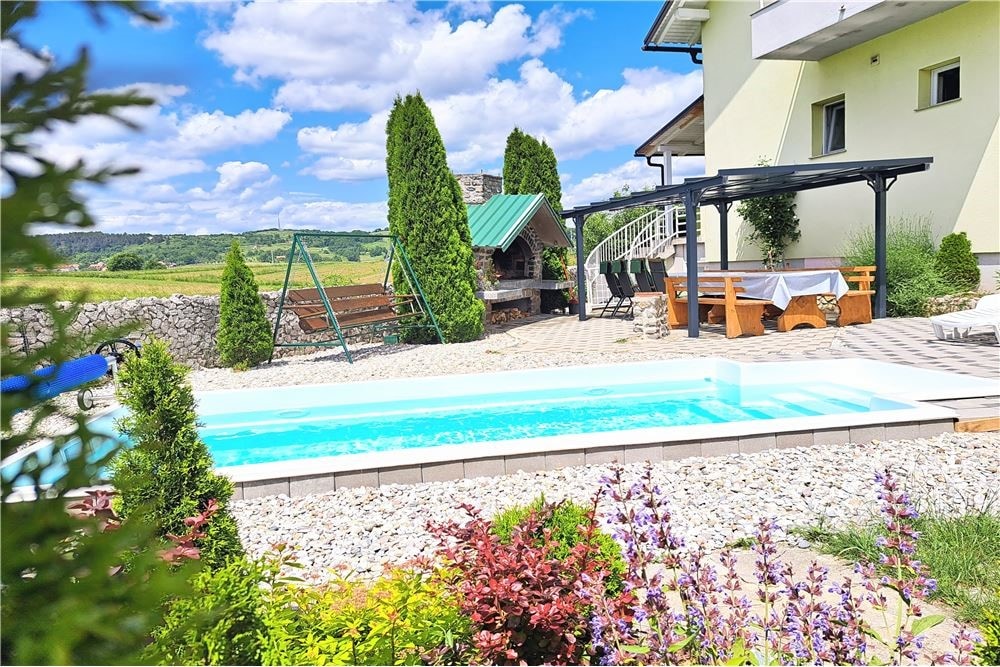 A private pool is surrounded by landscaped gardens, featuring a stone barbecue area and a shaded seating arrangement. Nearby, a swing set offers additional outdoor recreation. Lush greenery complements the tranquil countryside view, creating a peaceful atmosphere.