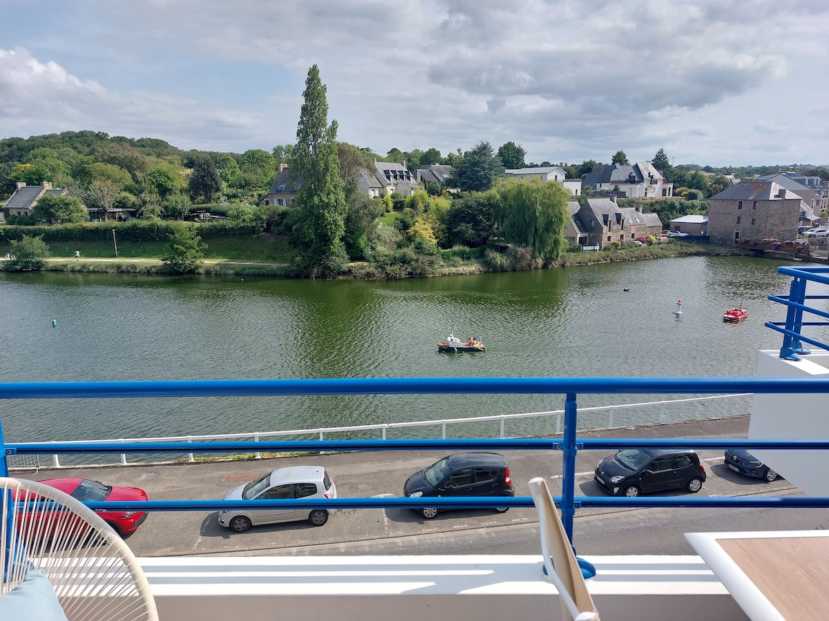 A view from the balcony overlooks a serene body of water flanked by lush greenery and residential buildings. A couple of small boats can be seen on the water, with parked cars lining the road below. The atmosphere appears peaceful and inviting.