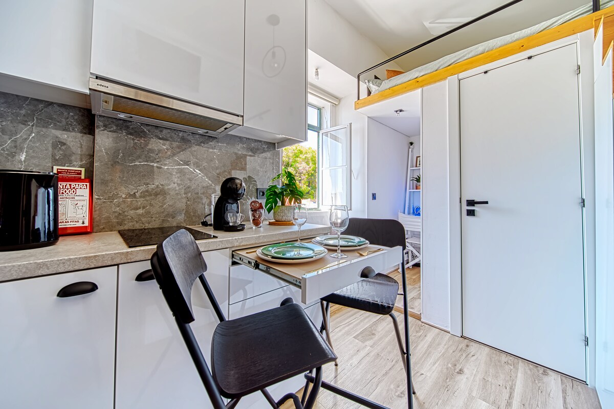 A compact kitchen area features modern appliances, including a coffee maker and a sleek stovetop. A table is set for two with dishware, accompanied by two black chairs. Light enters through a window, illuminating the contemporary cabinetry and decorative backsplash.