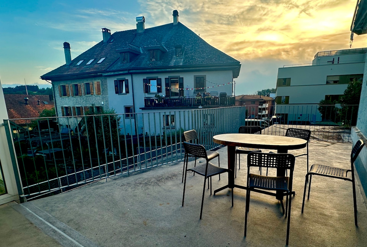 An outdoor terrace features a round table surrounded by four chairs, providing a space for gatherings. The backdrop showcases a charming building with multiple windows and a vibrant sunset sky, enhancing the outdoor experience.