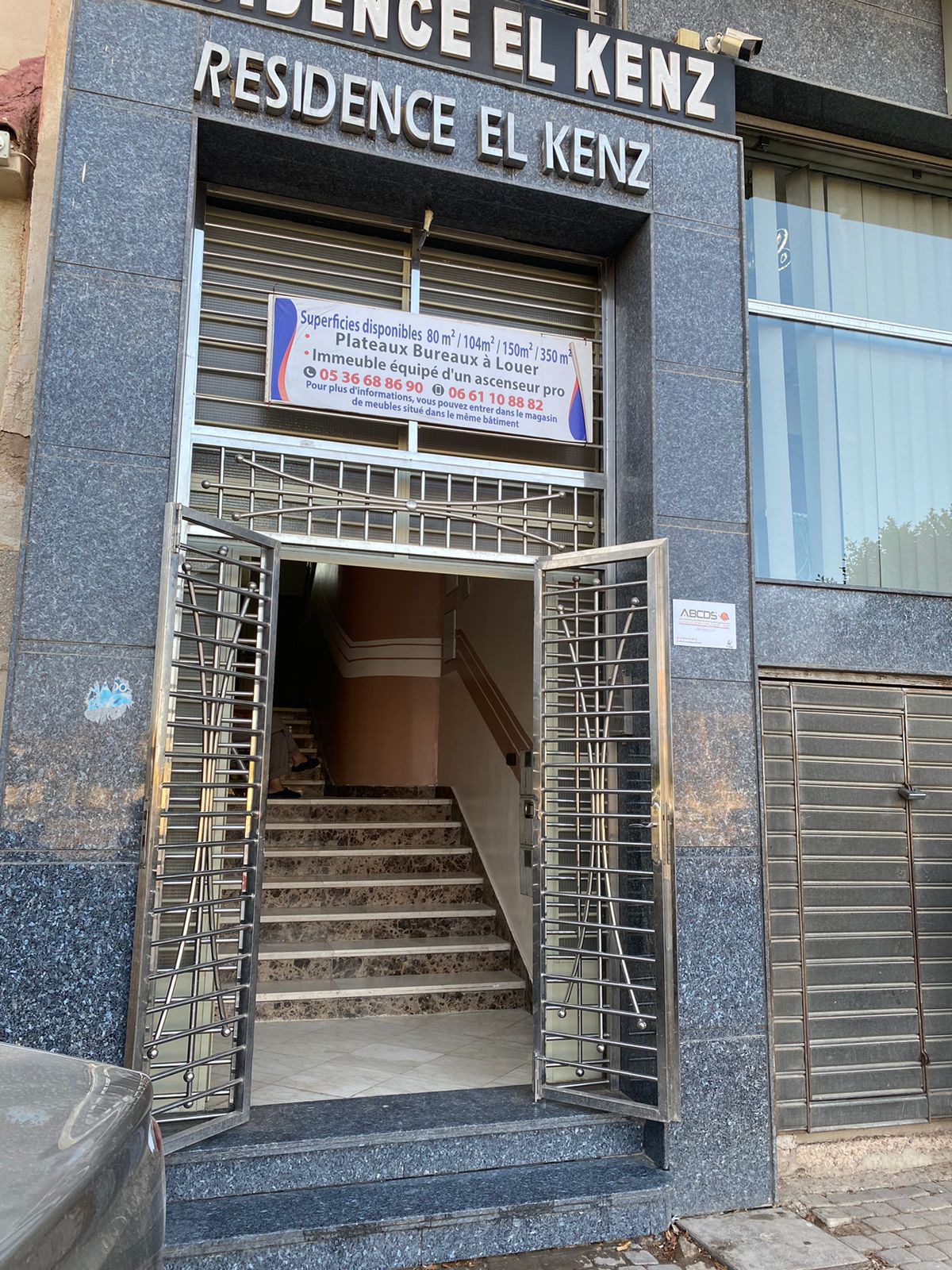 The building entrance features a sleek, modern design with a granite facade. Secure metal gates provide access to the interior, where a flight of stairs leads to the upper levels. A bright sign lists available rentals, enhancing visibility.