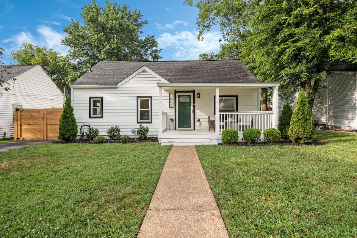 A charming bungalow is set amidst lush greenery, featuring a well-maintained front lawn. The white exterior is complemented by a welcoming porch with a green door. Flanking the entrance are small evergreen shrubs, enhancing the inviting appearance of the home.