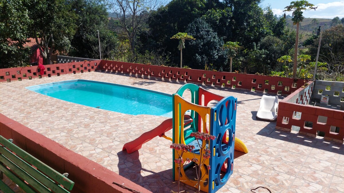 An outdoor area features a swimming pool surrounded by tiled flooring. A colorful children's play structure, including a slide and climbing area, is positioned nearby. Lush greenery and trees provide natural shade around the pool, creating a welcoming space for relaxation and play.
