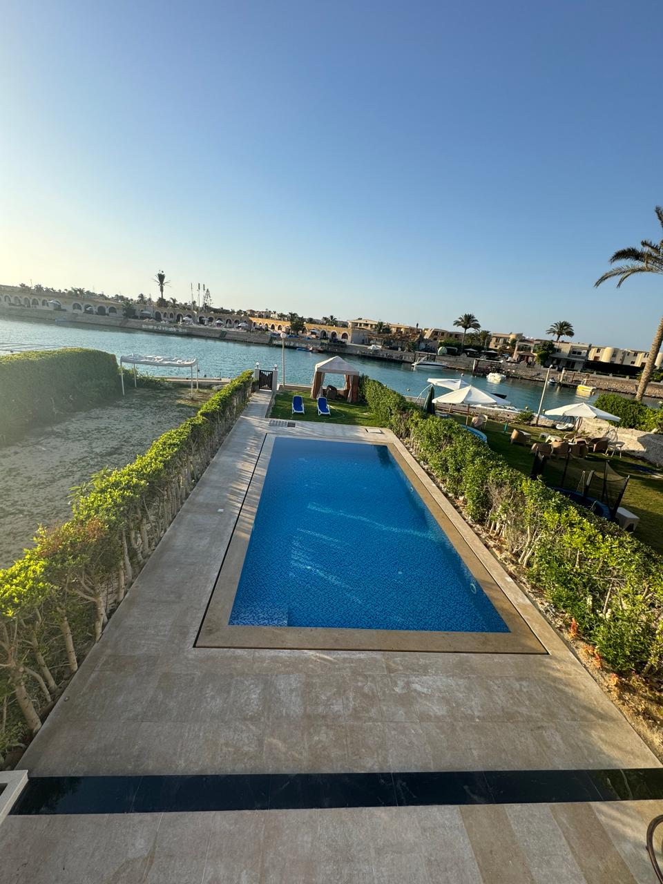 A private swimming pool is surrounded by neatly trimmed hedges and is set against a calm waterway. The pool area features sun loungers positioned on a stone patio, offering views of the lush greenery and nearby boats. A clear blue sky completes the serene outdoor setting.
