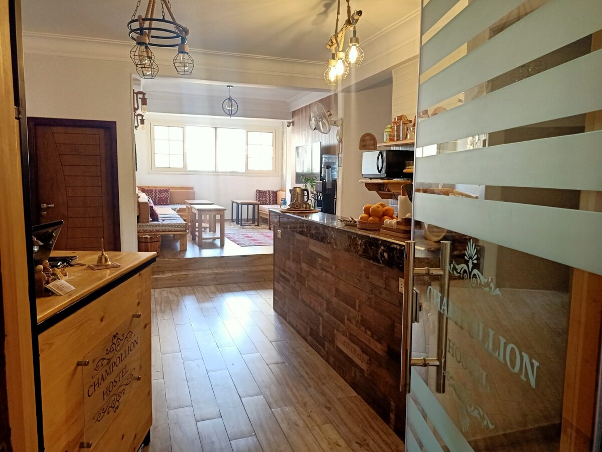 The shared kitchen is visible, featuring wooden cabinetry and a central island with a dark countertop. Natural light fills the space through large windows. Cozy seating with patterned cushions is arranged in the background, while decorative elements enhance the inviting ambiance.
