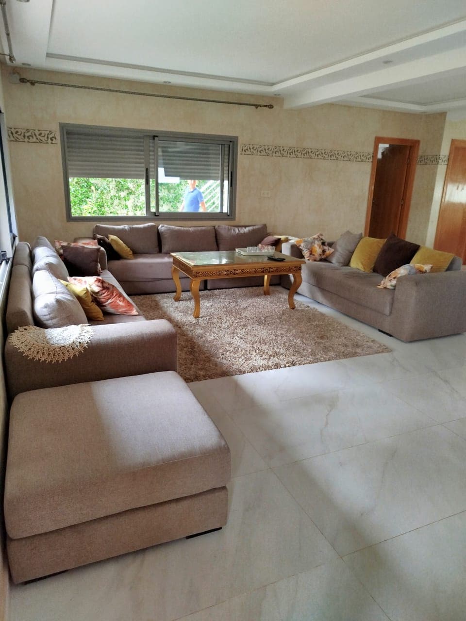 A spacious living room features a cozy arrangement of light-colored sofas with decorative pillows. A wooden coffee table sits atop a plush area rug, while large windows let in natural light, offering views of the outside greenery.