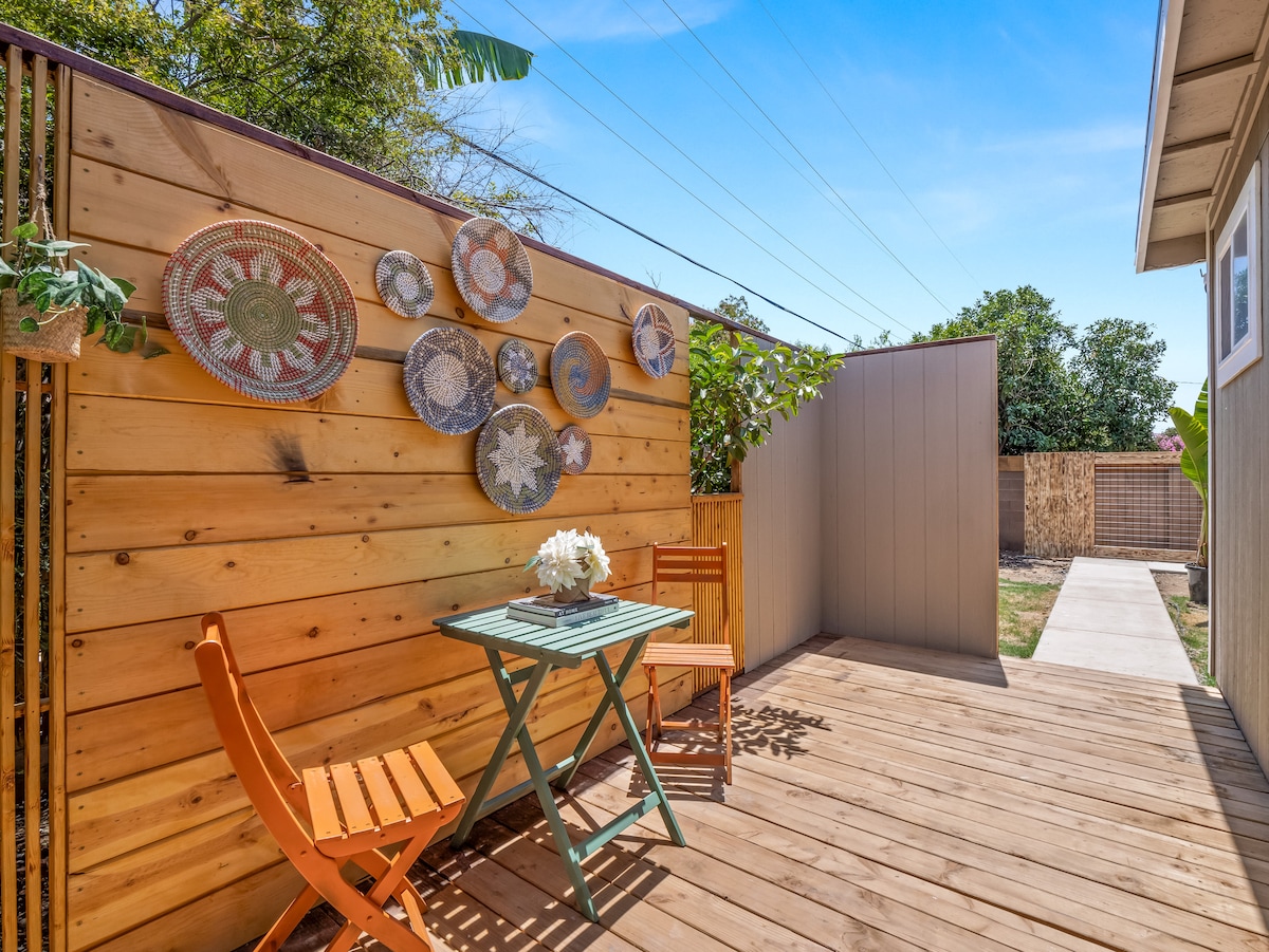 A wooden deck area is presented, featuring a small green table with a bouquet of flowers. Two orange chairs are positioned around the table. The backdrop consists of a wooden fence adorned with decorative woven wall art, complemented by greenery.