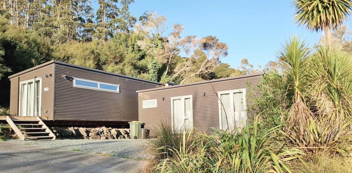 The exterior features two connected units with a modern design, set against a backdrop of greenery. Steps lead up to an expansive glass door, while large windows provide natural light. Lush plants and a gravel driveway surround the structure, enhancing the tranquil setting.