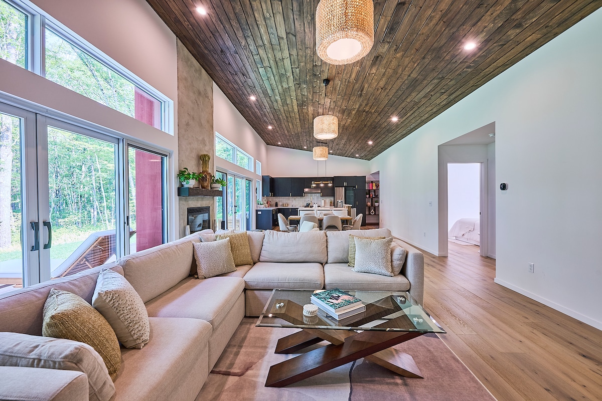 A spacious living area features a large L-shaped couch adorned with decorative pillows, accompanied by a modern coffee table. Bright natural light streams through expansive glass doors, illuminating the wooden ceiling with elegant pendant lights. A dining area in the background offers a glimpse of the kitchen appliances.