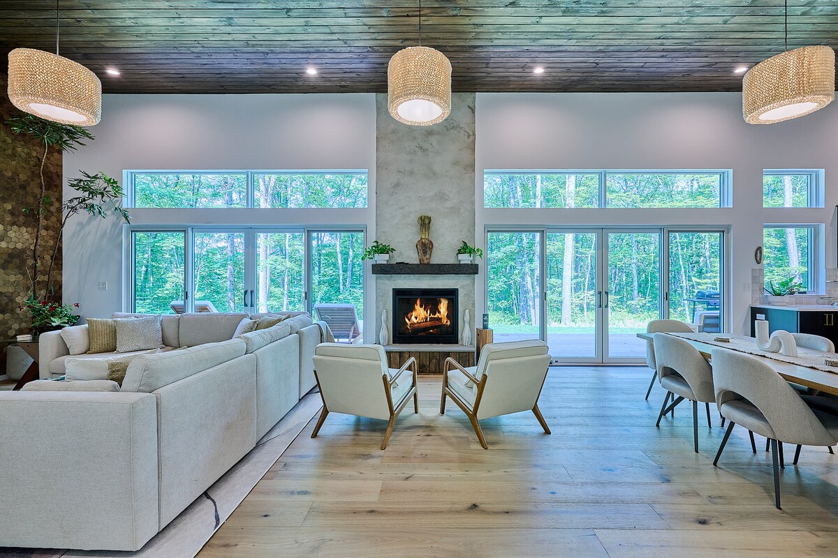 The spacious living area features a large L-shaped couch facing a cozy fireplace, with two comfortable armchairs positioned nearby. Expansive windows provide natural light and forest views, while pendant lighting enhances the modern aesthetic of the space.