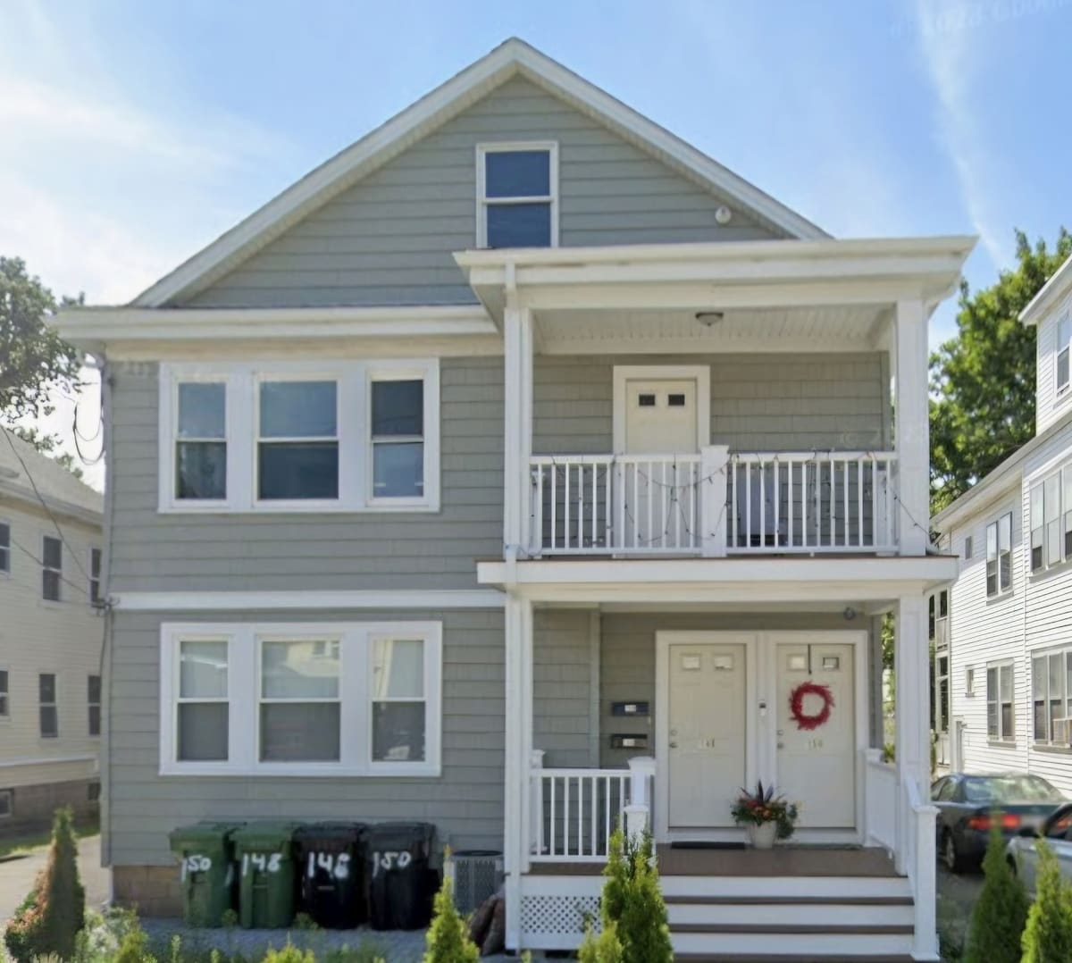 An exterior view of a two-story house is presented, featuring a light gray facade with white trim. A welcoming front porch is visible, adorned with a holiday wreath. Decorative shrubs line the entrance, and green trash bins are positioned in front of the property.