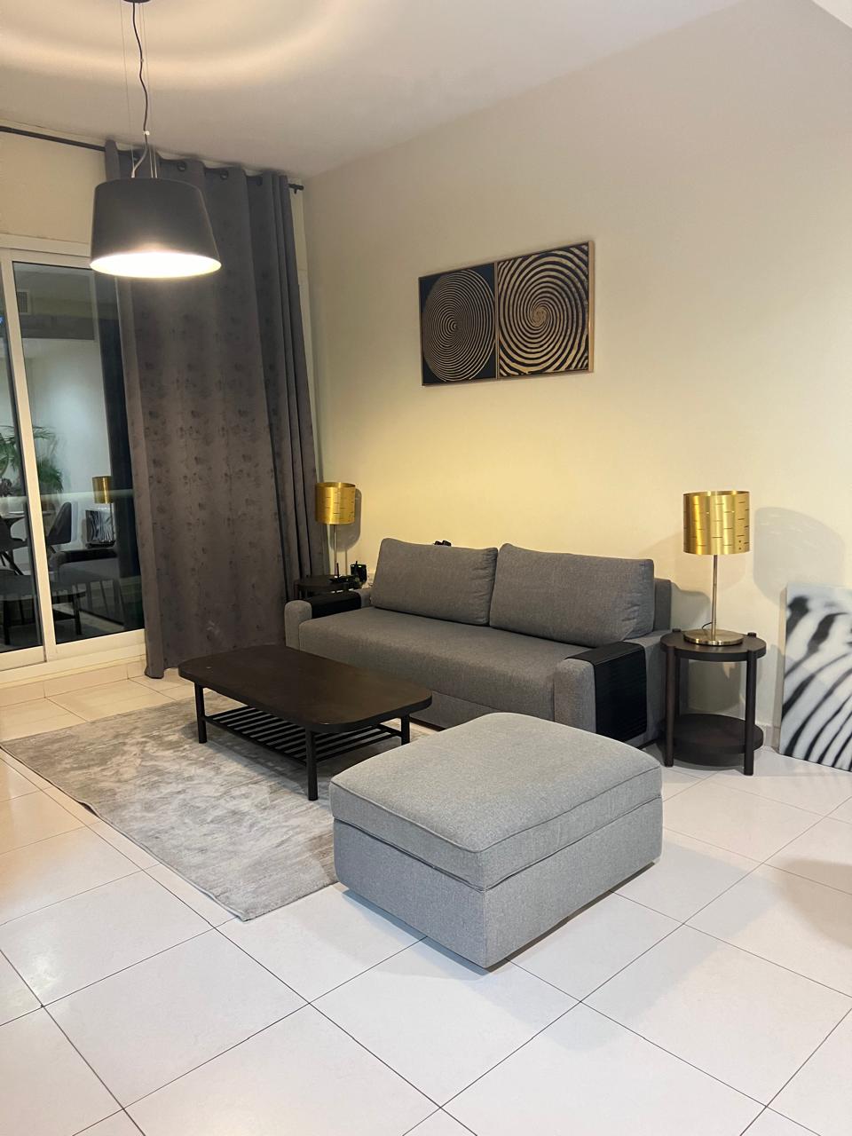 A comfortable living area features a gray sofa and an ottoman, complemented by a coffee table. Gold table lamps flank the seating arrangement, and a circular rug adds warmth to the tiled floor. Floor-to-ceiling curtains frame the large windows, enhancing the spacious feel.