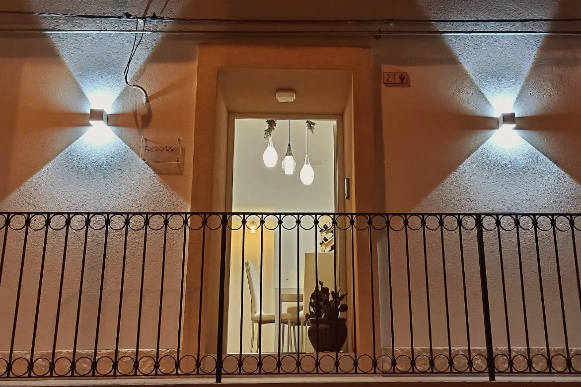 The entrance features warm lighting with two wall-mounted lights illuminating the doorway. A decorative plant sits on the balcony railing. Inside, a dining table and chairs are visible through the open door, blending modern design with a welcoming ambiance.