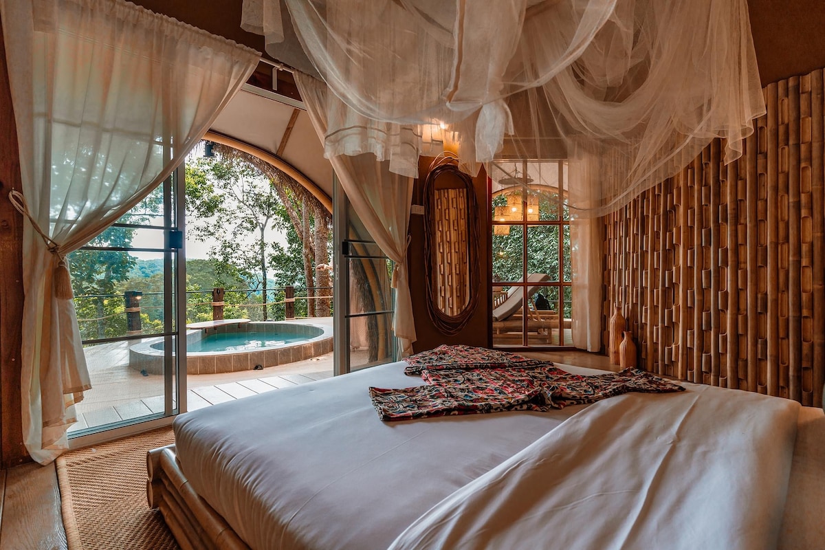 A spacious guest room features a large bed with a light-colored duvet, complemented by a decorative textile. Sheer curtains allow natural light to fill the room. A private terrace shows a view of an outdoor hot tub surrounded by greenery.