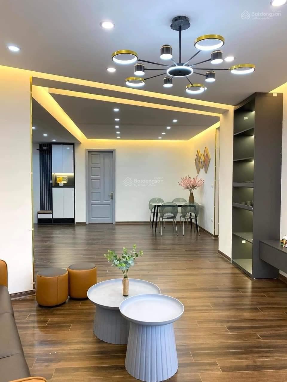 A modern living area is illuminated by stylish recessed lighting. A comfortable seating arrangement is complemented by two round coffee tables. A dining space with a glass table and four chairs is visible, alongside a gray door and sleek shelving against wooden flooring.
