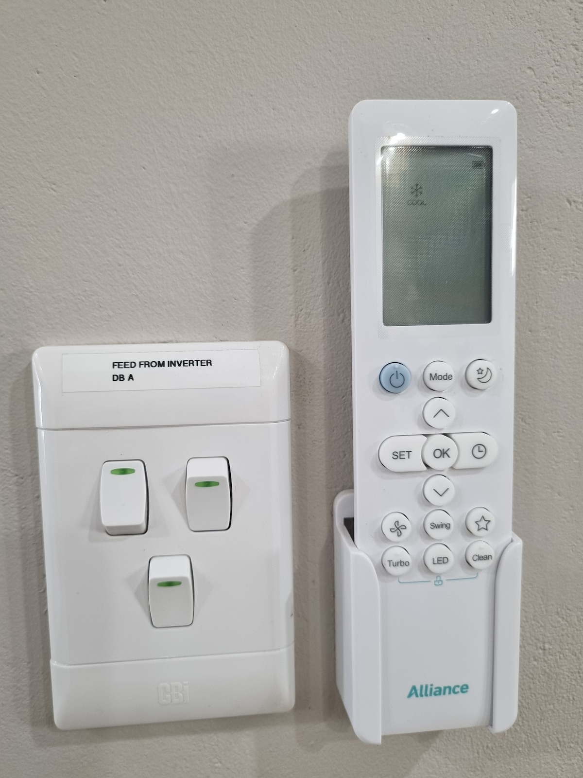 An air conditioning remote is housed in a wall-mounted holder next to a power switch panel. The panel features two switches, each with green indicators, indicating functionality. The remote control is clearly labeled with buttons for temperature adjustment and various modes.