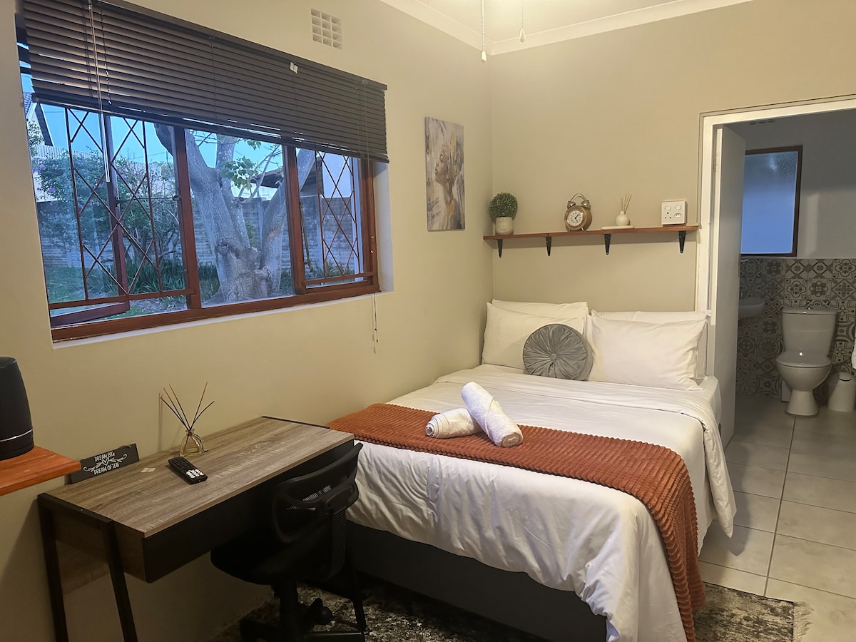 A cozy sleeping area features a spacious bed with light linens and an orange throw. A small desk is positioned nearby, and natural light filters through a window adorned with dark blinds. A minimalist shelf displays decorative items, and a bathroom entrance is visible in the background.