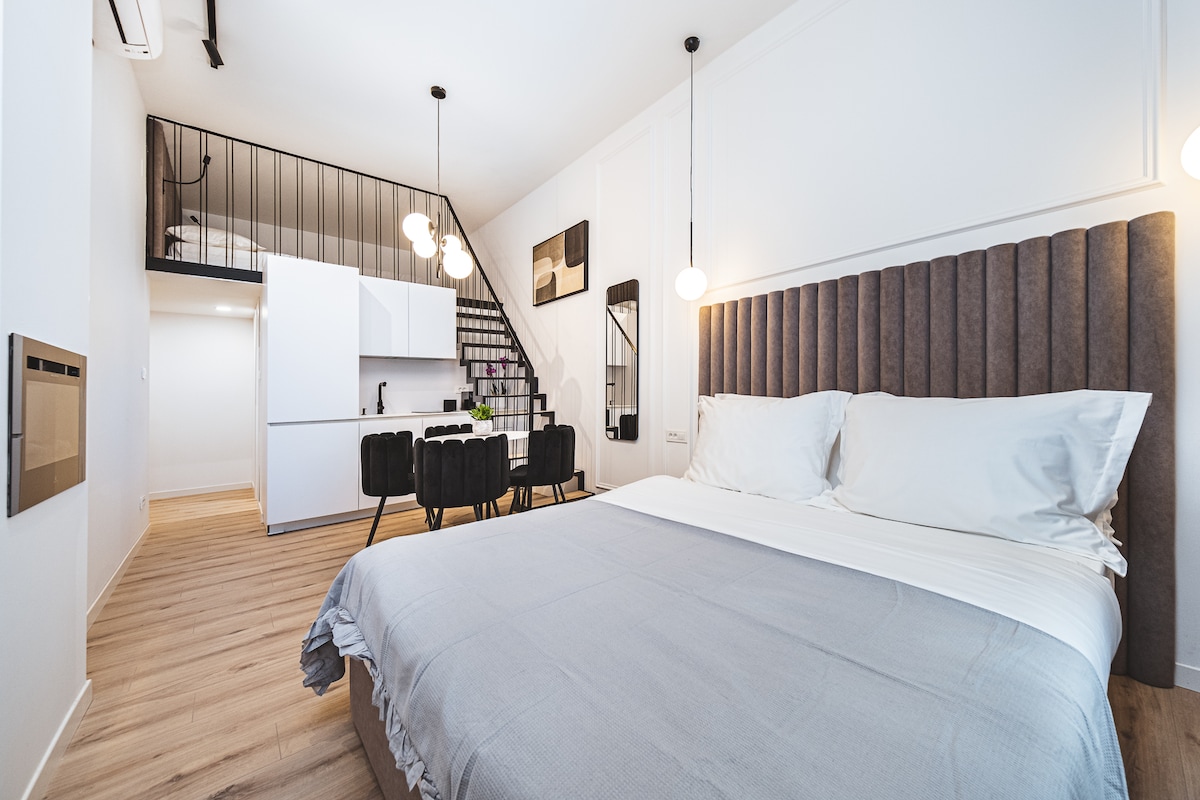 A spacious sleeping area features a large bed with a soft grey blanket. In the background, a staircase leads to an upper level. A compact dining area with four black chairs is visible beside a modern kitchen cabinetry, completing the inviting layout.